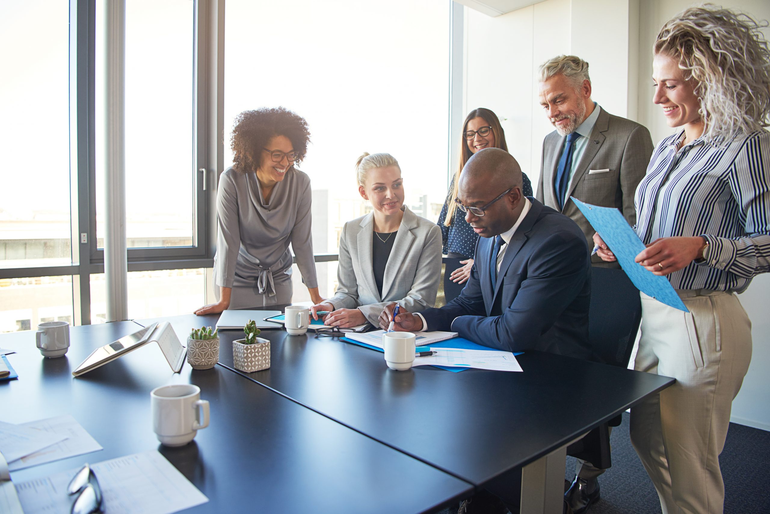 Diverse group of businesspeople standing around their manager sitting at an office boardroom table reviewing charts and paperwork